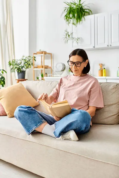 Eine reife Frau in bequemer Homewear sitzt auf einer Couch und ist ganz in ein Buch vertieft. — Stockfoto