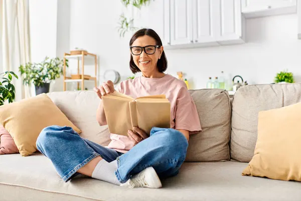 A mature woman in cozy homewear relaxes on a couch, engrossed in reading a book. — Stock Photo