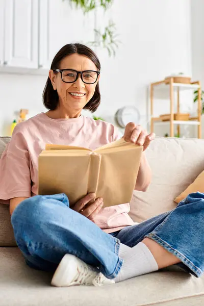 Зріла жінка в затишному домашньому одязі, сидячи на дивані, захоплена читанням книги. — стокове фото