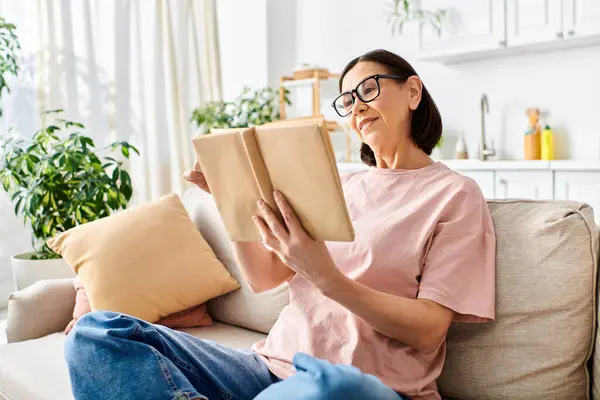 Reife Frau in Homewear, auf dem Sofa sitzend, in ein Buch vertieft. — Stockfoto