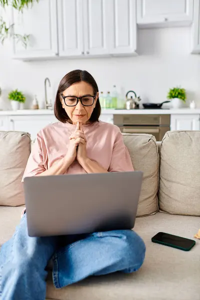 Una mujer madura en ropa de casa acogedora se sienta en un sofá, escribiendo en un ordenador portátil. - foto de stock