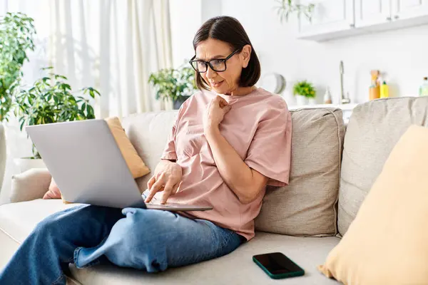 Reife Frau in gemütlicher Kleidung sitzt auf einer Couch, konzentriert auf einem Laptop-Bildschirm. — Stockfoto