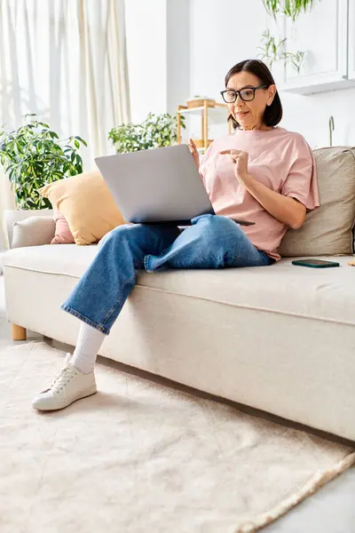 A mature woman in cozy homewear sitting on a couch, using a laptop. — Stock Photo