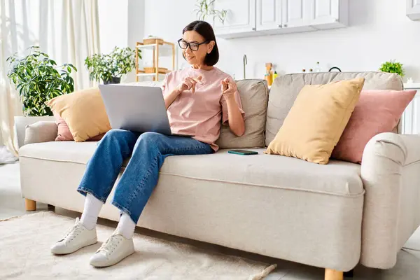 Una mujer en ropa de casa acogedora se sienta en un sofá, absorbida en su computadora portátil. - foto de stock