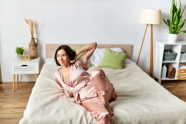 A mature woman dressed in pink pajamas is relaxing on a bed in a cozy home setting. — Stock Photo