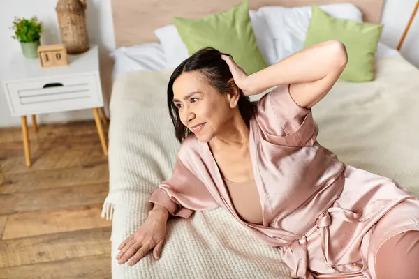 A mature woman in a pink dress sits gracefully on a bed in a cozy setting. — Stock Photo