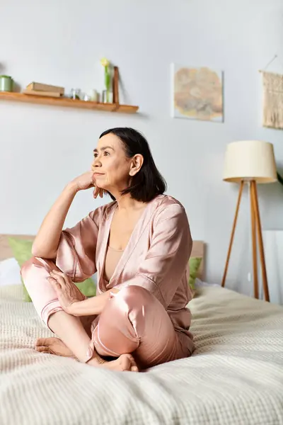 A mature woman in cozy homewear sits peacefully on a bed next to a glowing lamp. — Stock Photo