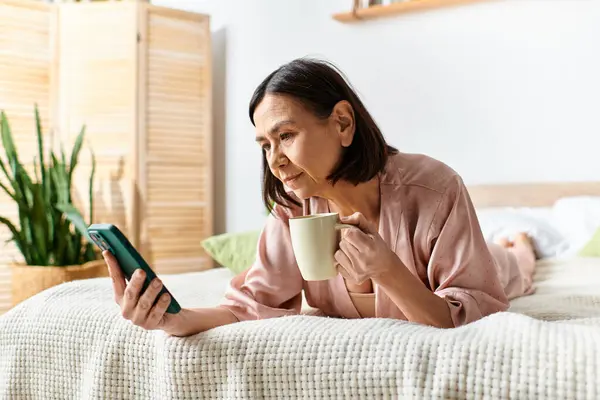 Una mujer en acogedora ropa de casa se relaja en una cama, sosteniendo una taza de café y mirando su teléfono. - foto de stock