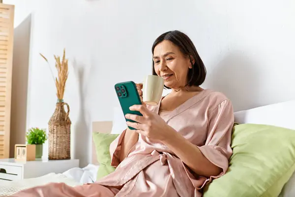 Una donna in accogliente biancheria da casa seduta su un letto, assorta nel suo telefono cellulare. — Stock Photo