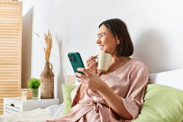 Woman in cozy homewear enjoys coffee while checking phone on bed. — Stock Photo