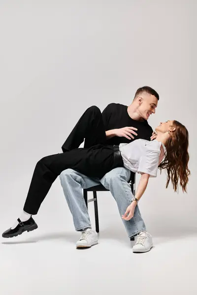 Un giovane uomo e una giovane donna si siedono insieme su una sedia, condividendo un tenero momento di amore e connessione su uno sfondo grigio. — Foto stock