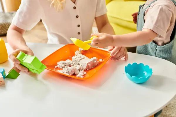 A toddler girl joyfully explores a plate of sand while her mother watches closely in a Montessori-style learning activity. — Stock Photo