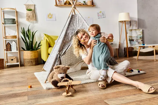 A curly-haired mother and her toddler daughter sit on the floor in front of a teepee tent, engaging in the Montessori method of education at home. — Stock Photo