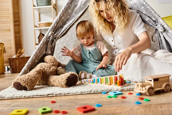 Lockige Mutter und Kleinkind-Tochter beim fantasievollen Spielen in einem bunten Spielzelt im gemütlichen Heim-Ambiente. — Stockfoto