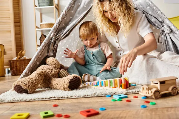 Curly mother and toddler daughter engaging in imaginative play inside a colorful play tent in a cozy home setting. — Stock Photo