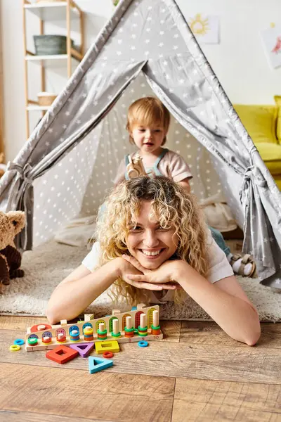 Eine lockige Mutter liegt mit ihrer kleinen Tochter auf dem Boden und erkundet Montessori-Aktivitäten zu Hause. — Stockfoto