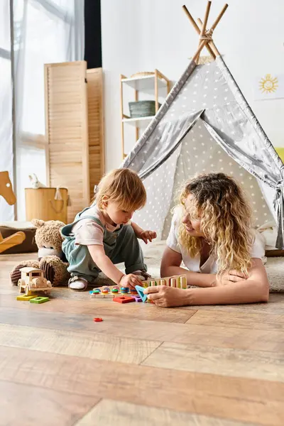 Una madre dai capelli ricci e sua figlia bambina giocano felicemente con i giocattoli sul pavimento, praticando il metodo Montessori di educazione.. — Foto stock