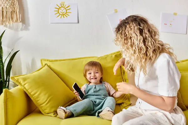 A curly-haired mother lovingly combs her toddler daughters hair while sitting on a cozy yellow couch at home. — Stock Photo