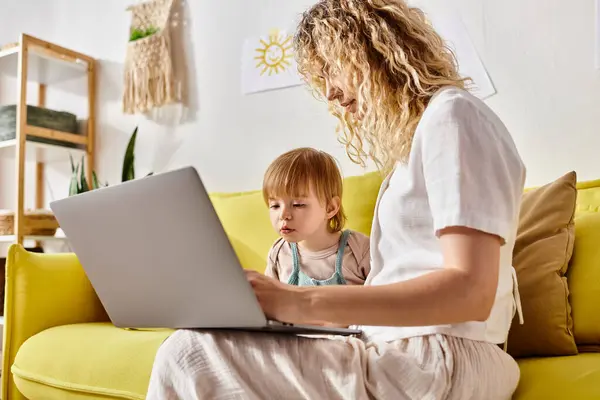 A curly-haired mother sits on a couch with her toddler daughter on her laptop, bonding and sharing a moment of love and affection. — Stock Photo