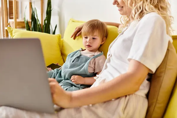 A curly-haired mother sitting on a couch, tenderly holding her toddler daughter on her lap in a cozy home setting. — Stock Photo