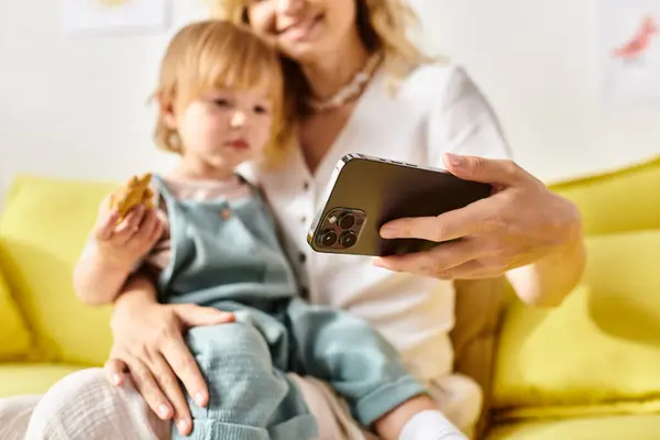 A curly mother holds a cell phone next to her toddler daughter at home, engaging in a heartwarming moment of connection. — Stock Photo