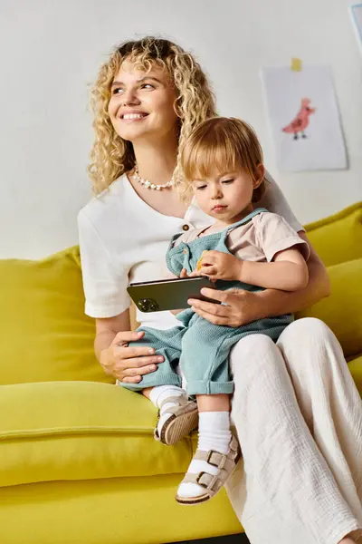 Eine lockige Mutter sitzt auf einer Couch und umarmt ihre kleine Tochter zu Hause liebevoll. — Stockfoto