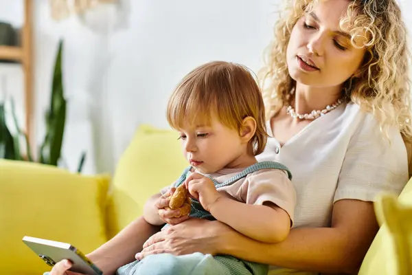 A curly-haired mother lovingly holds her toddler daughter while sitting on a cozy couch at home, screen time — Stock Photo
