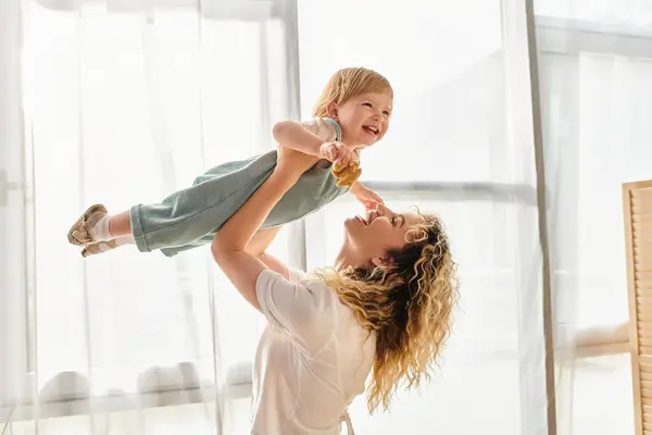 Eine lockige Mutter hebt freudig ihre kleine Tochter in die Luft und drückt damit Liebe und Verspieltheit zu Hause aus. — Stockfoto