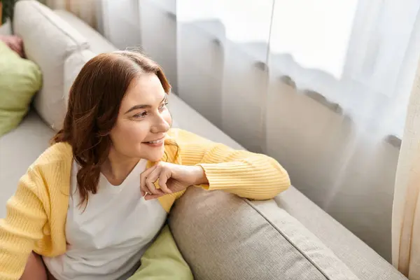 Middle aged woman in contemplation, perched on couch by a window. — Stock Photo