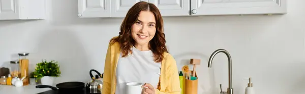 Middle-aged woman standing in kitchen, holding cup. — Stock Photo