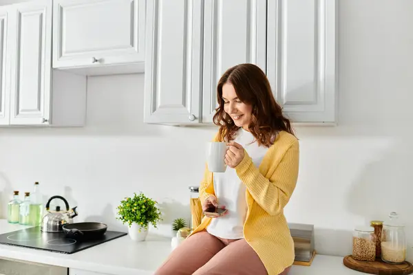 Женщина средних лет наслаждается моментом расслабления, держа чашку, сидя на кухонном столе. — стоковое фото