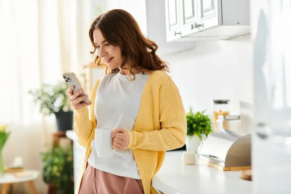 A middle-aged woman standing in a kitchen, absorbed in her cell phone. — Stock Photo