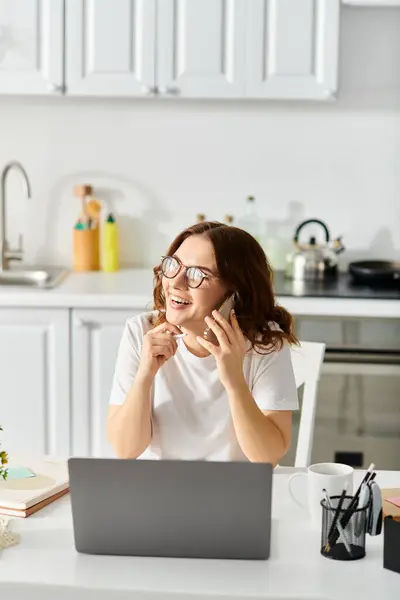 A middle-aged woman sits at a kitchen table, engrossed in a conversation on her cell phone. — Stock Photo