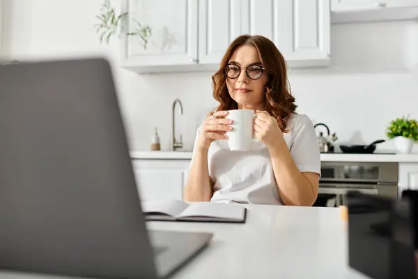 Una mujer de mediana edad se sienta en una mesa con un portátil y una taza de café. - foto de stock