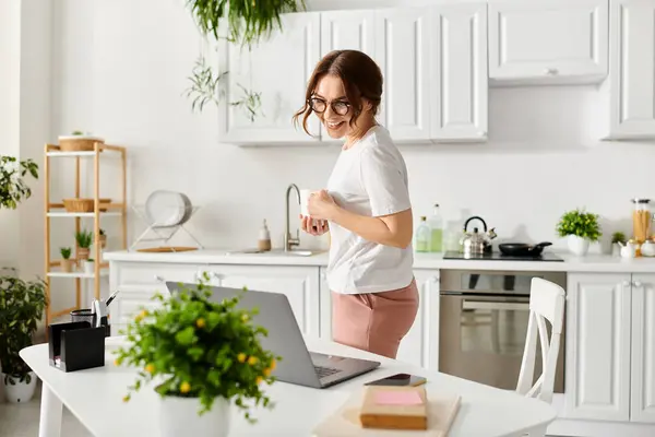 Middle-aged woman standing on kitchen counter, multitasking with laptop. — Stock Photo