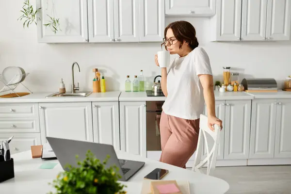 Una mujer de mediana edad se encuentra en una cocina junto a un ordenador portátil, comprometida con una tarea digital. - foto de stock