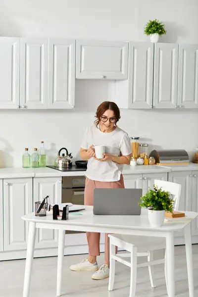 Middle aged beauty stands in kitchen, laptop in hand. — Stock Photo