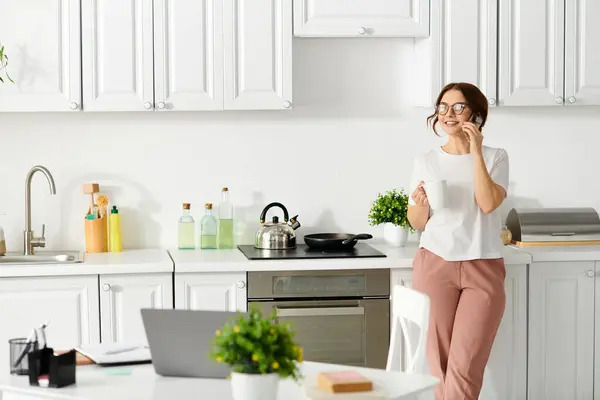 Middle-aged woman standing in a kitchen, talking on a cell phone. — Stock Photo