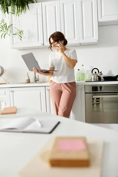 Middle-aged woman stands in kitchen, holding a laptop. — Stock Photo
