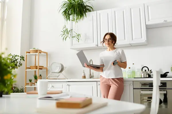 A middle-aged woman stands in the kitchen, holding a laptop. — Stock Photo