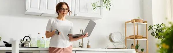 Uma mulher de meia-idade fica em sua cozinha segurando um laptop, misturando tecnologia e cozinhando. — Fotografia de Stock