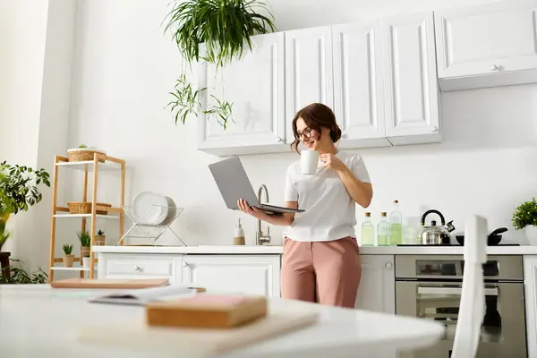 Middle-aged woman standing in kitchen, multitasking with laptop in hand. — Stock Photo