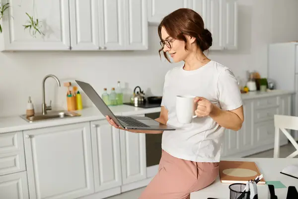 Middle aged woman relaxing at home, holding coffee cup and working on laptop. — Stock Photo