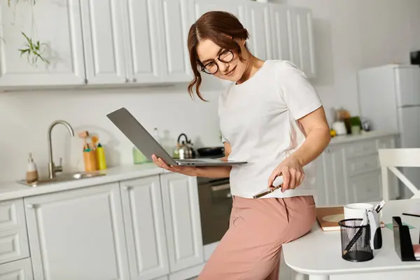Frau mittleren Alters steht mit Laptop auf Küchentisch. — Stockfoto