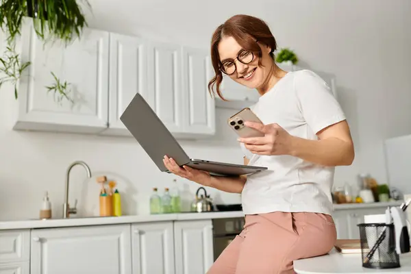 Middle-aged woman sits on counter, typing on laptop. — Stockfoto