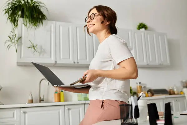 Mulher de meia-idade multitarefa na cozinha, segurando um laptop. — Fotografia de Stock