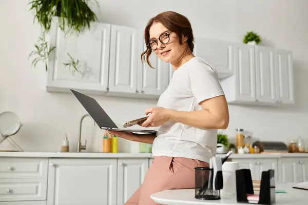 Middle aged woman sitting on counter, using laptop. — Stock Photo