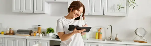 Middle-aged woman standing and engrossed in reading a book in a kitchen. — Stock Photo