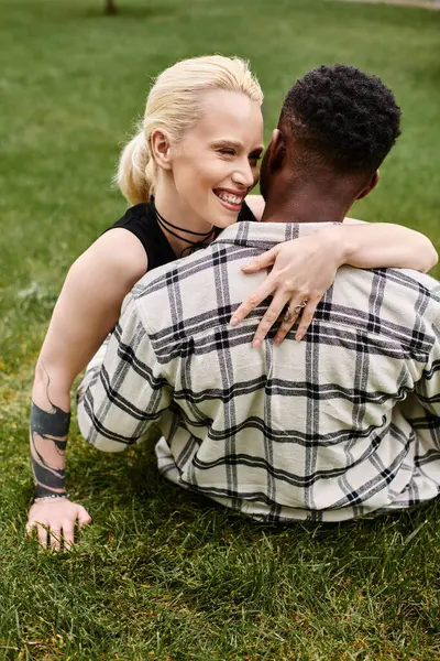 Una feliz pareja multicultural, un hombre afroamericano y una mujer caucásica, compartiendo un tierno abrazo en la hierba en un parque. - foto de stock