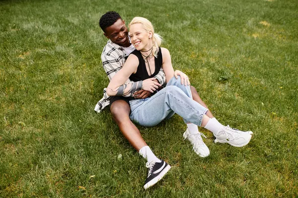 Un hombre afroamericano feliz y una mujer caucásica sentados pacíficamente juntos en la hierba verde en un parque. - foto de stock